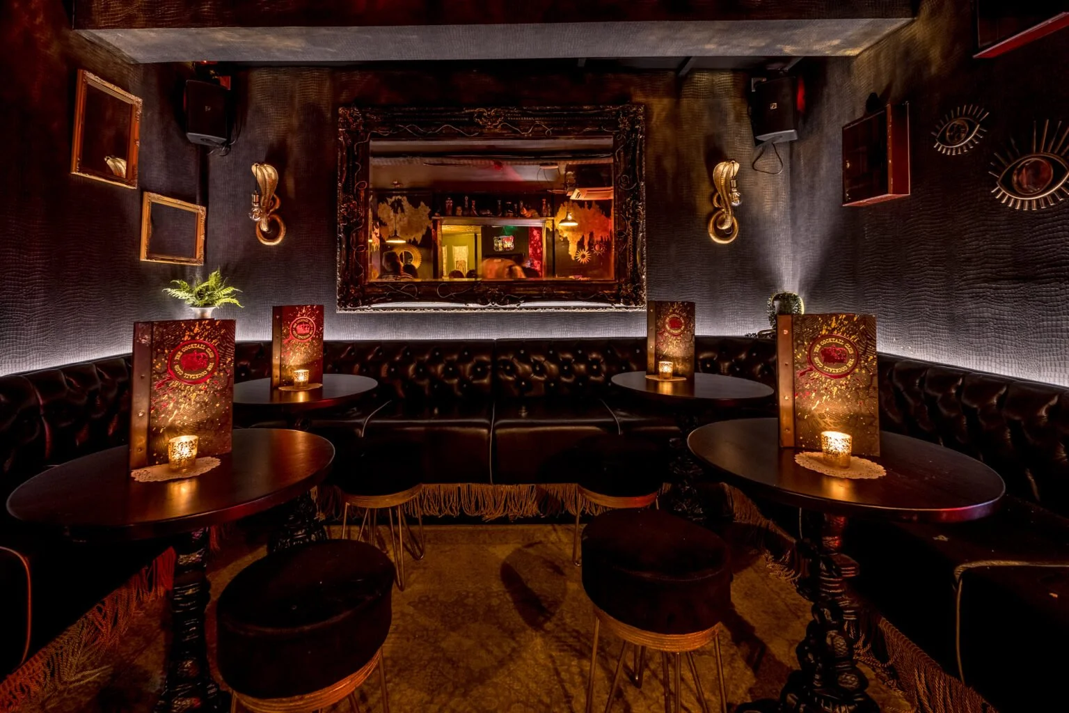 Dimly lit lounge with black leather seating and small round tables, candles, and decorative mirrors and artwork on dark textured walls.