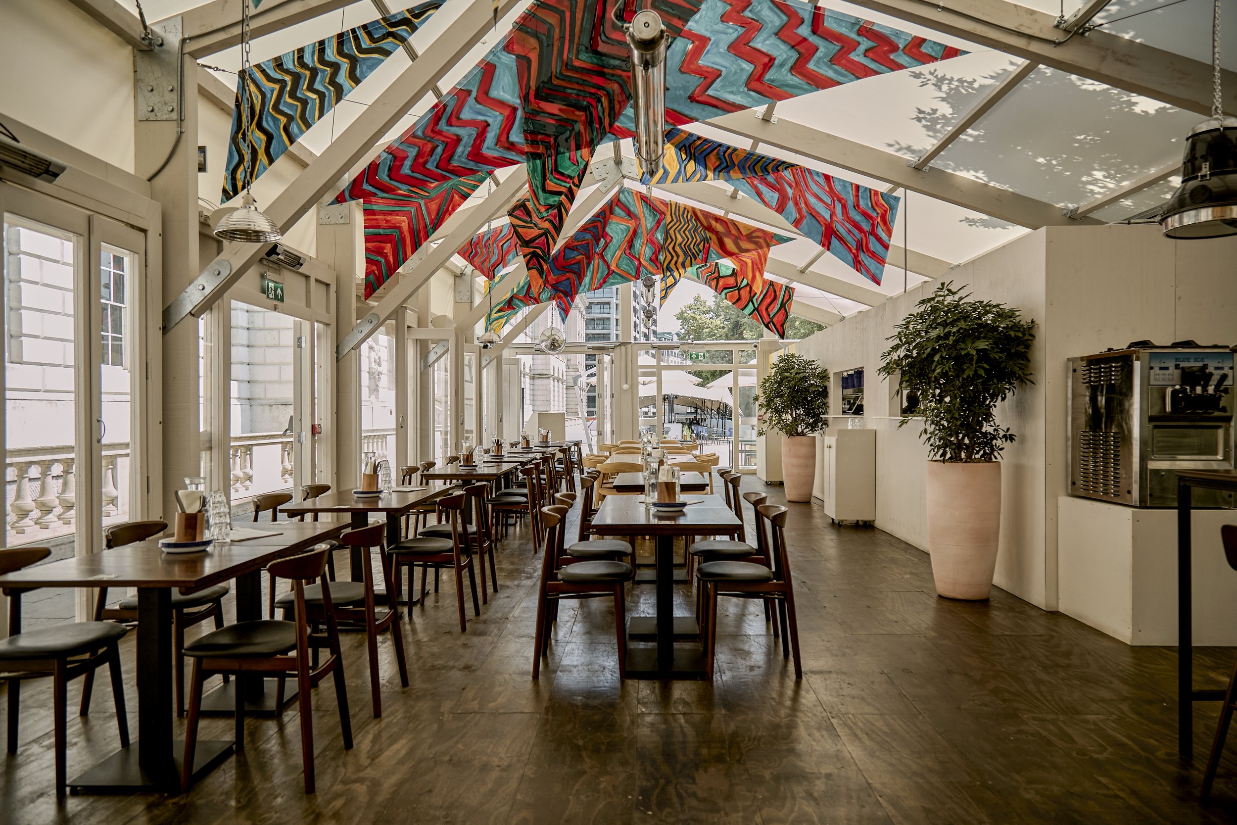 Interior of a bright, modern restaurant with wooden floors, large windows, colorful patterned fabric banners hanging from the ceiling, and potted plants along the wall.