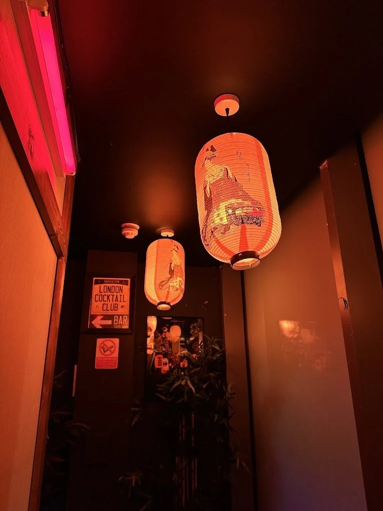 Three orange Japanese-style paper lanterns hanging from a dark ceiling, with a neon pink sign, a 'London Cocktail Club' sign, and a plant in the background.