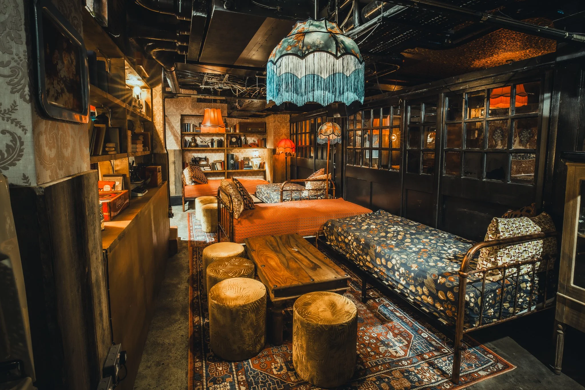 A cozy cafe or lounge with vintage decor, including multiple beds or daybeds with patterned cushions, a wooden coffee table, plush stools, hanging lamps with fabric shades, shelves with books, and a dark ceiling with exposed pipes.