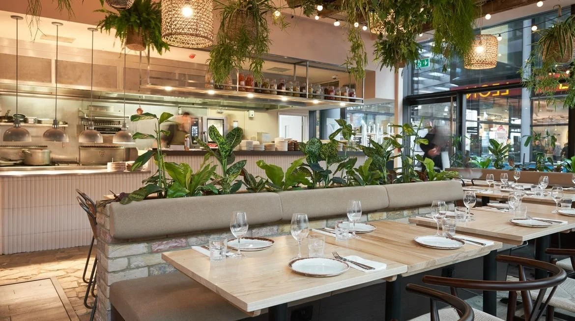 Inside a modern restaurant with wooden tables set for dining, surrounded by green plants and hanging greenery, with open kitchen visible in background.