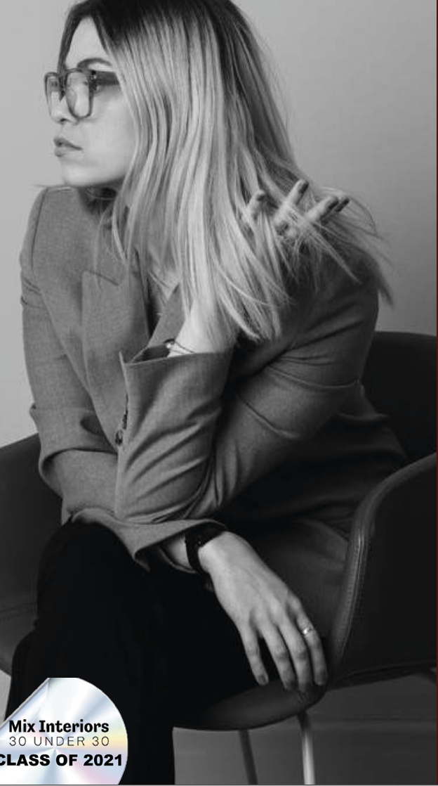 A woman with shoulder-length hair wearing glasses and a suit, sitting on a chair, resting her chin on her hand, in a black and white photograph.