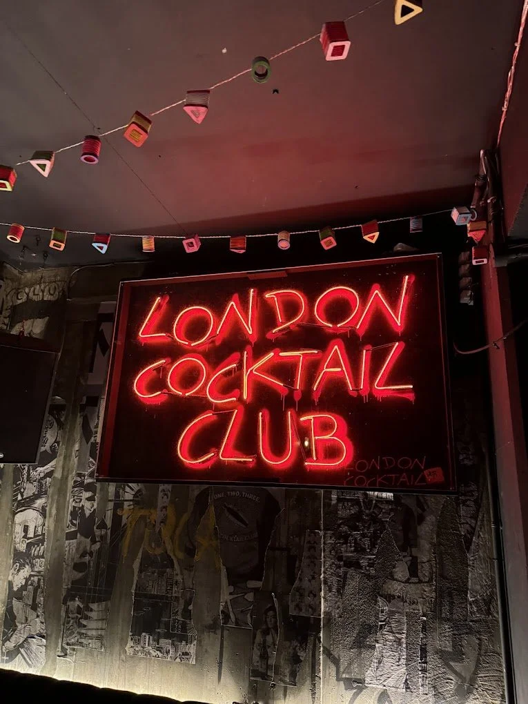 Red neon sign that reads "London Cocktail Club" against a wall with black and white collage artwork, with colorful hanging decorations above.
