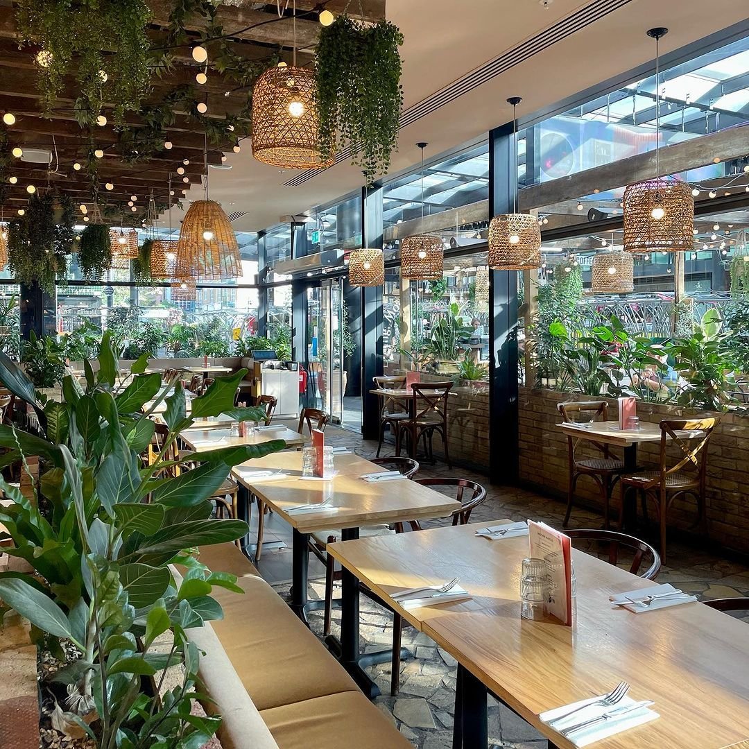 Interior of a cozy restaurant with wooden tables, chairs, and abundant greenery. Decor includes hanging woven lamps, string lights, and plants hanging from the ceiling, with large glass windows letting in natural light.