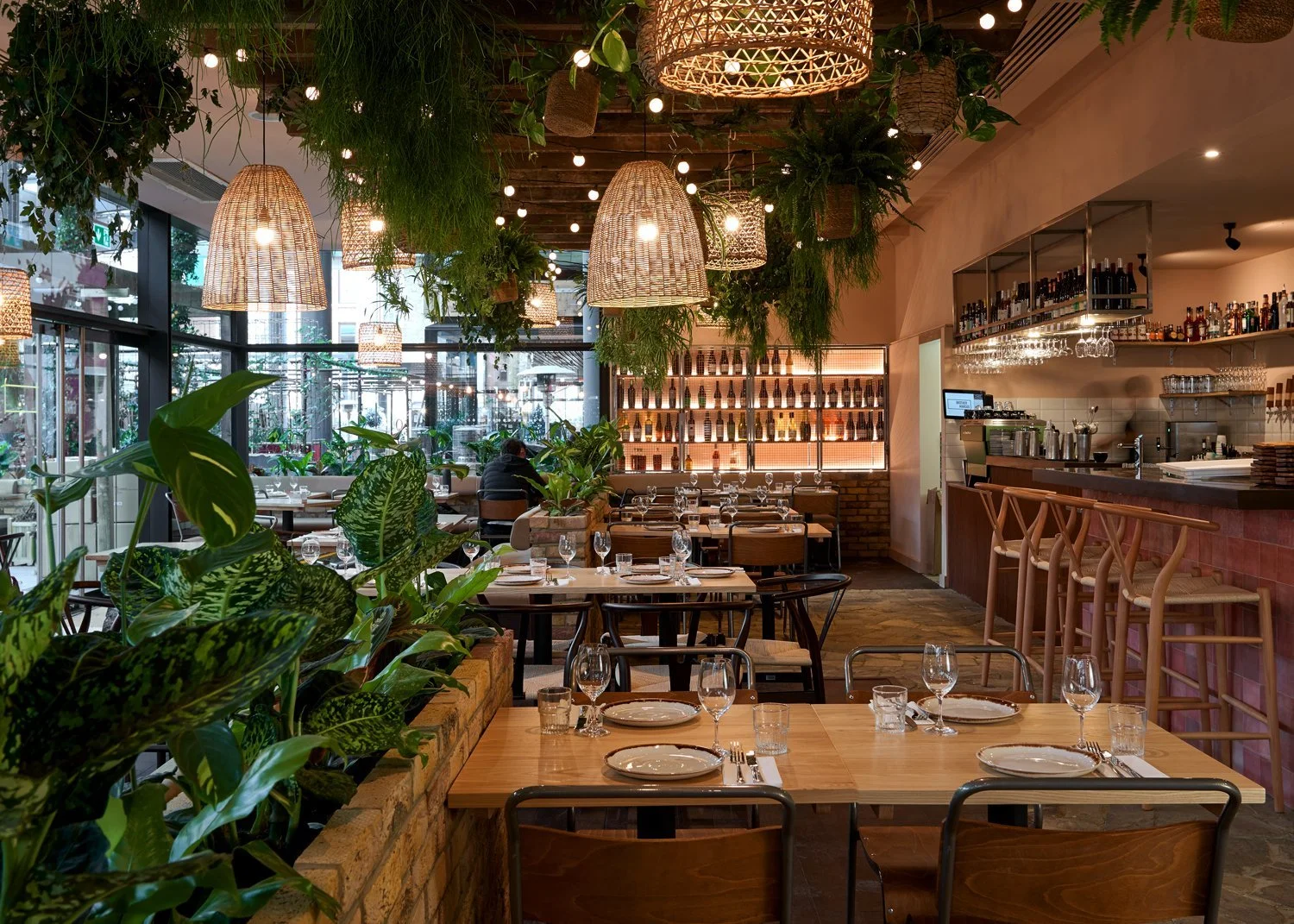 Interior of a modern restaurant with wooden tables set for dining, surrounded by green plants, large hanging wicker lamps, and a bar area with barstools and shelves of bottles.