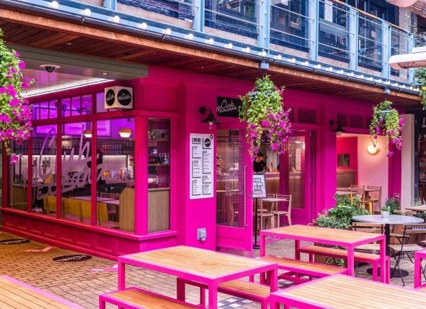 A vibrant pink vegan fast casual restaurant with outdoor seating, large glass windows, hanging flower baskets, and purple lighting inside.
