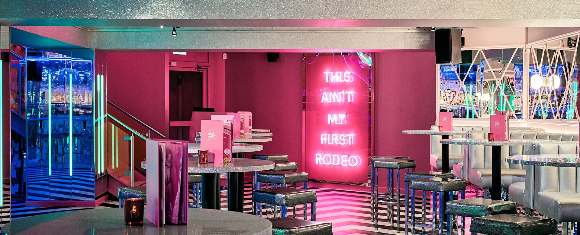 Colorful neon-lit interior of a restaurant or bar with seating, tables, and a pink wall with neon sign that says "THIS AIN'T MY FIRST RODEO."
