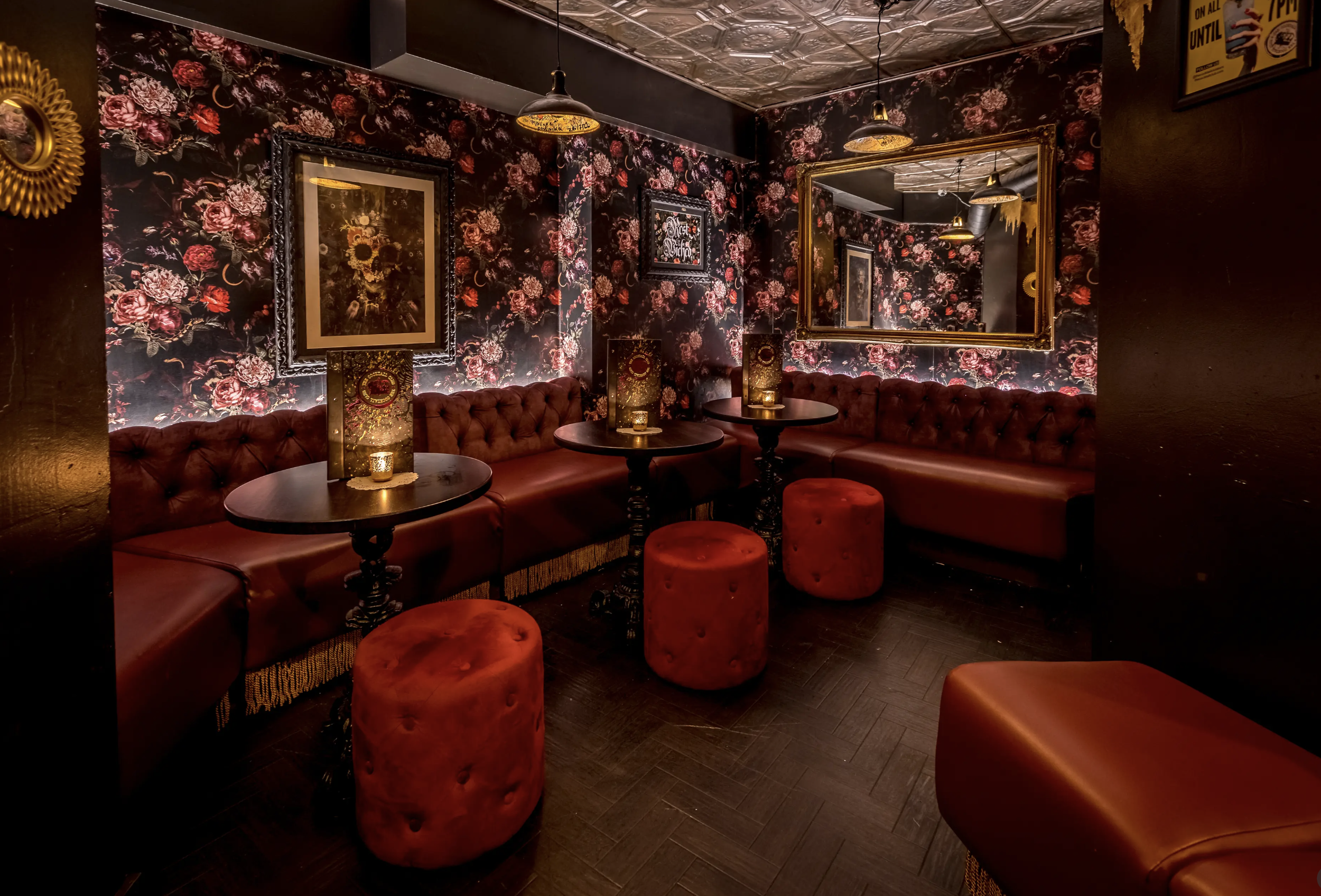 A cozy corner of a dimly lit bar with red leather tufted banquettes, dark wooden tables, red velvet stools, floral wallpaper, framed artwork, a large mirror, and hanging pendant lights.
