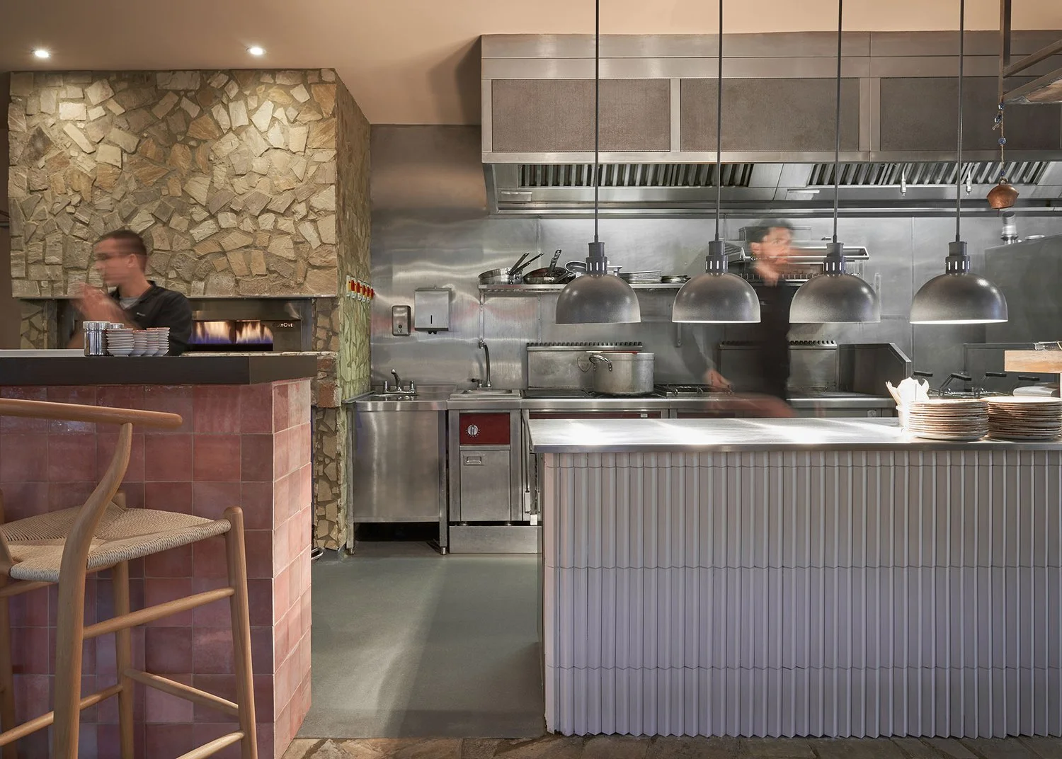 Interior of a modern restaurant kitchen with two chefs, stainless steel appliances, and a stone wall.