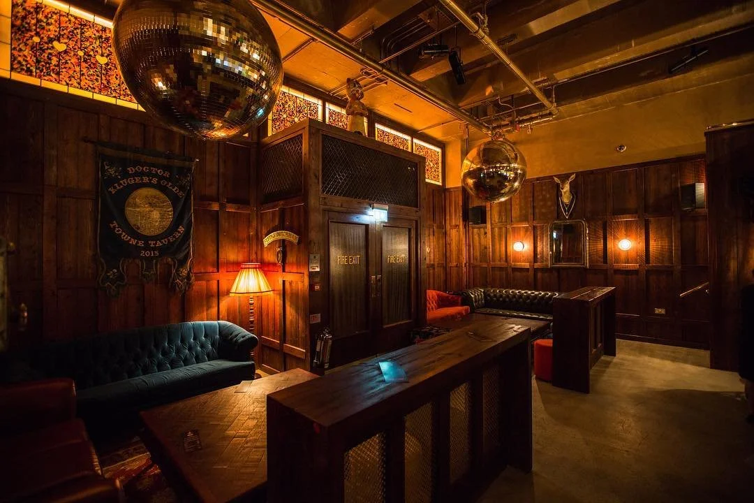 A cozy dimly lit lounge with dark wood paneling, a green tufted sofa, orange chairs, a wooden table, and disco balls hanging from the ceiling.