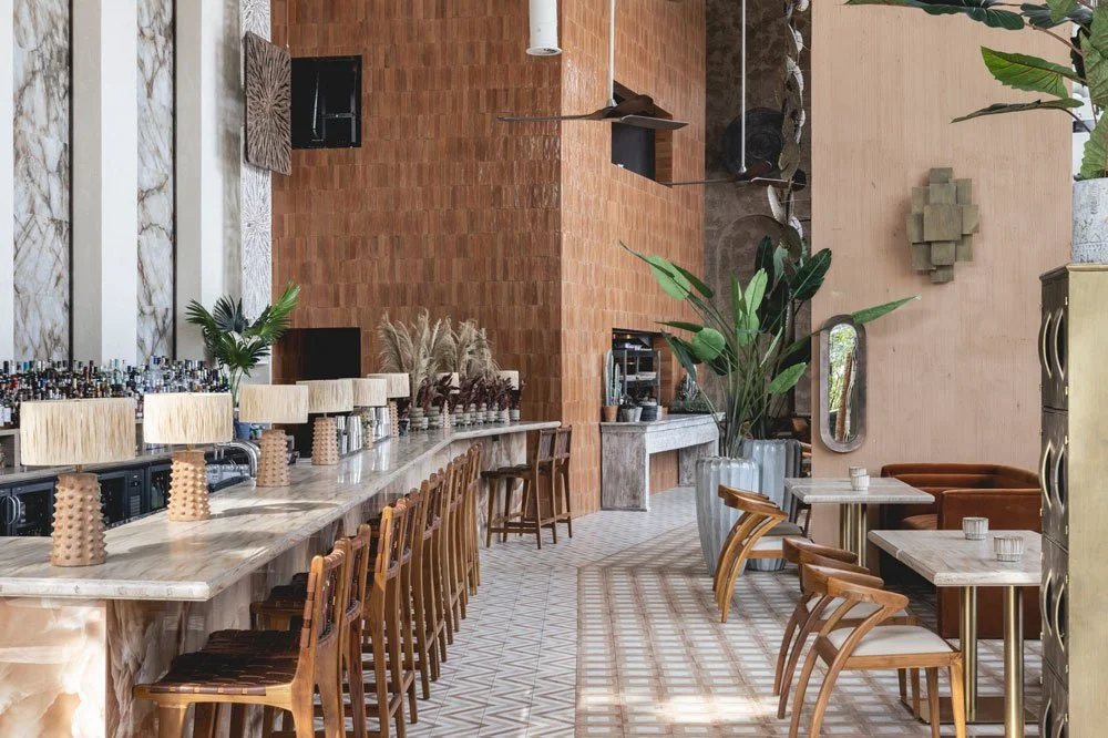 Interior of a stylish cafe or restaurant with a marble bar counter, wooden chairs, and potted plants, featuring warm-toned walls and natural light.