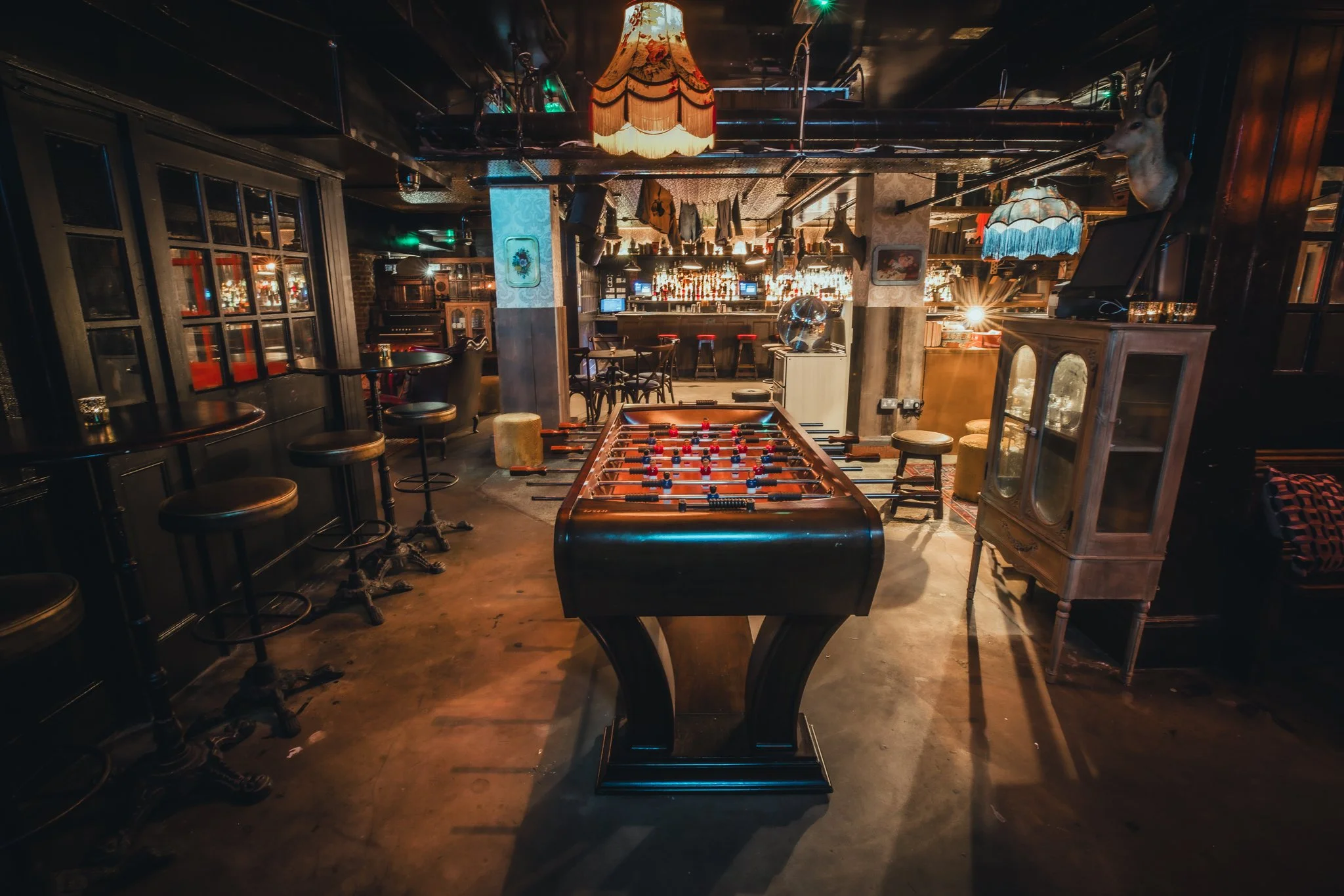Interior of a bar with a foosball table in the center, high tables with bar stools on the sides, decorative lamps, a bar counter at the back, and mounted animal head on the wall.