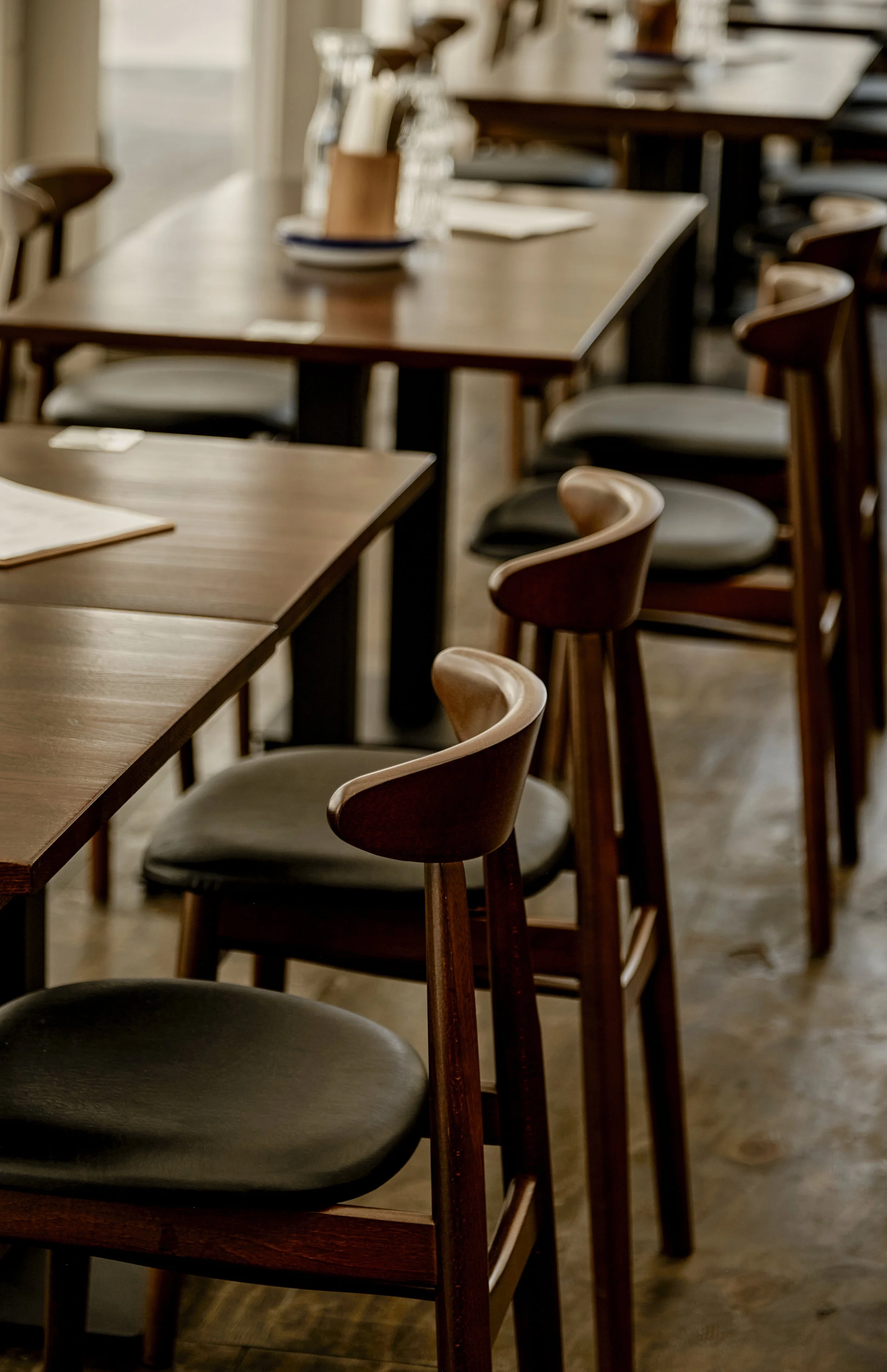 Interior view of a restaurant with wooden tables and chairs, some chairs with black cushioned seats, and table settings with plates and utensils.