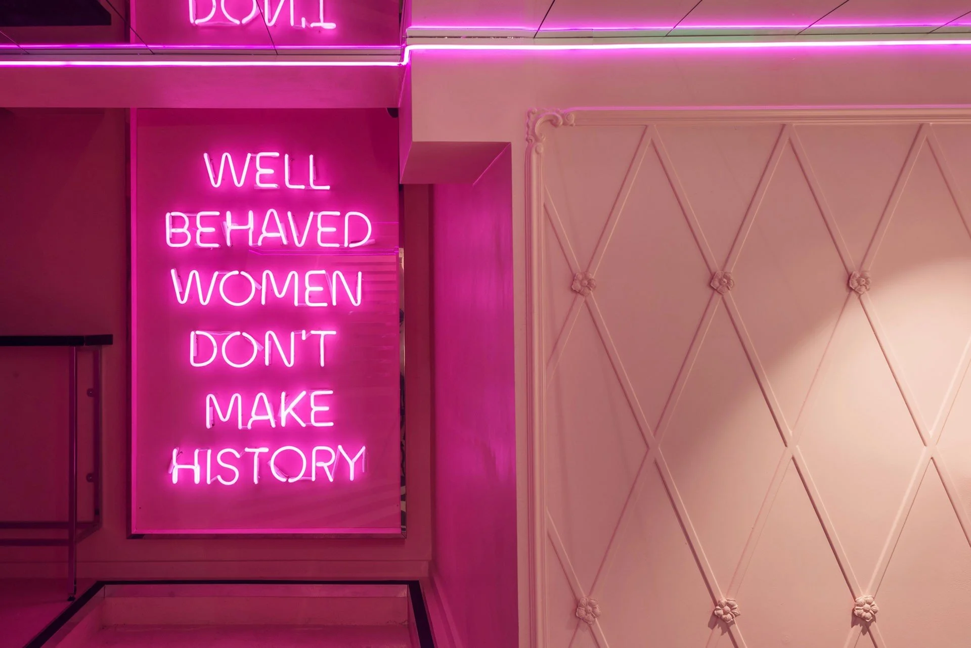 Pink neon sign with the text 'Well Behaved Women Don't Make History' in a modern interior with pink lighting and wall decor.