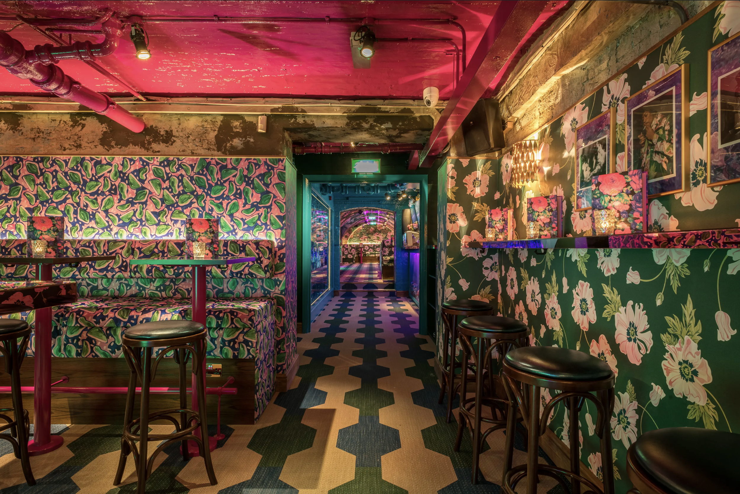 Interior of a colorful bar with floral and fruit patterned wallpaper, bar stools, and framed photos, illuminated with pink and blue lighting.