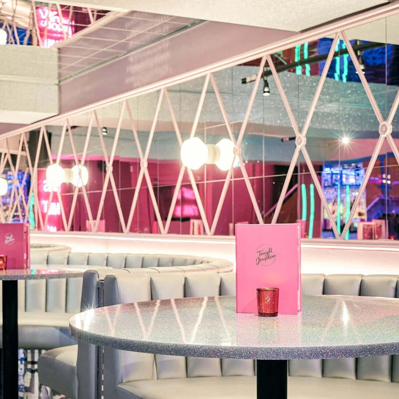 A round table with a pink menu and red candle holder, surrounded by white cushioned booths, in a modern, neon-lit restaurant with pink and neon signage reflected in the mirror.