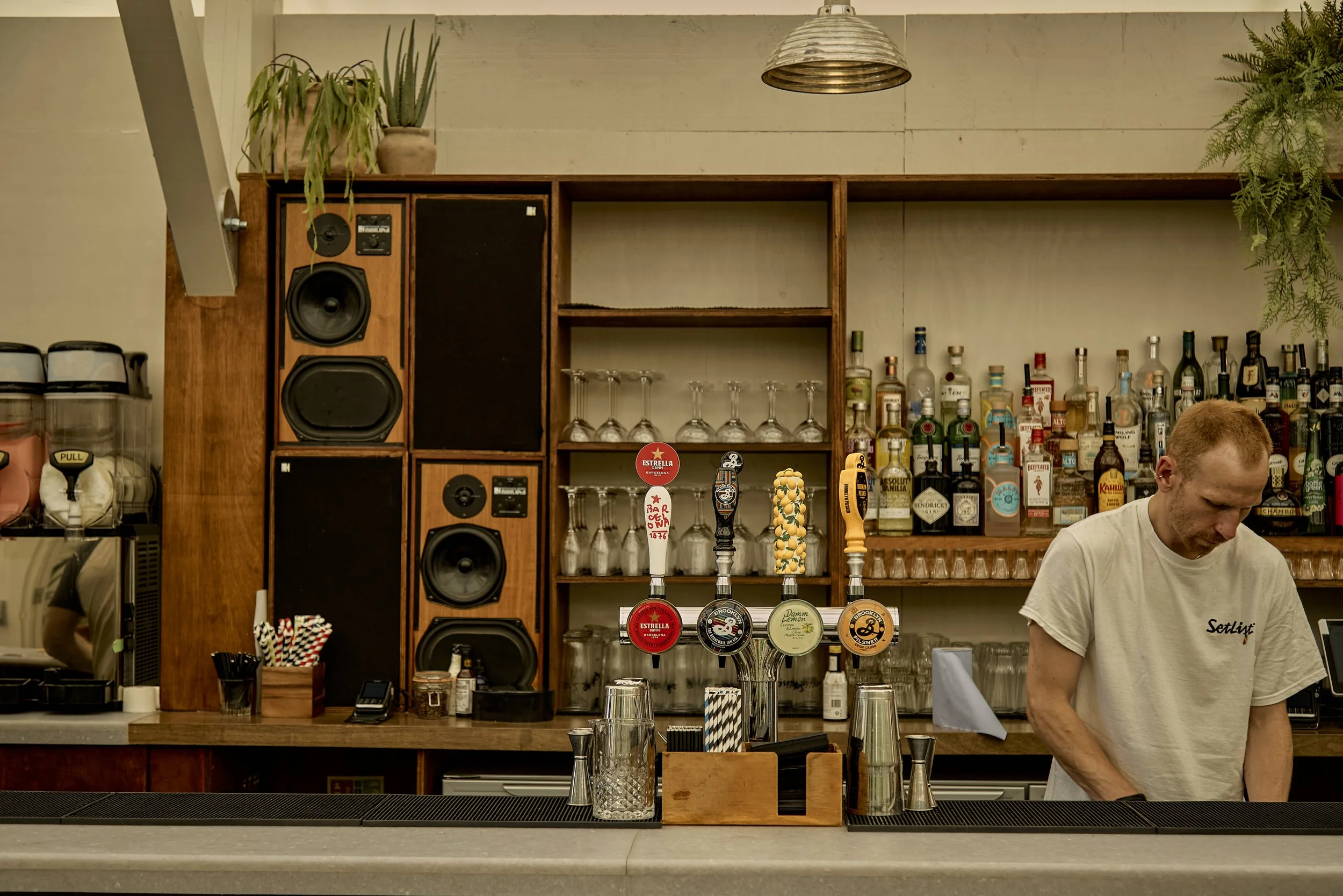 A bartender with orange hair working behind a bar with liquor bottles in the background. Four beer taps are visible on the counter, with various glasses and bar tools around.