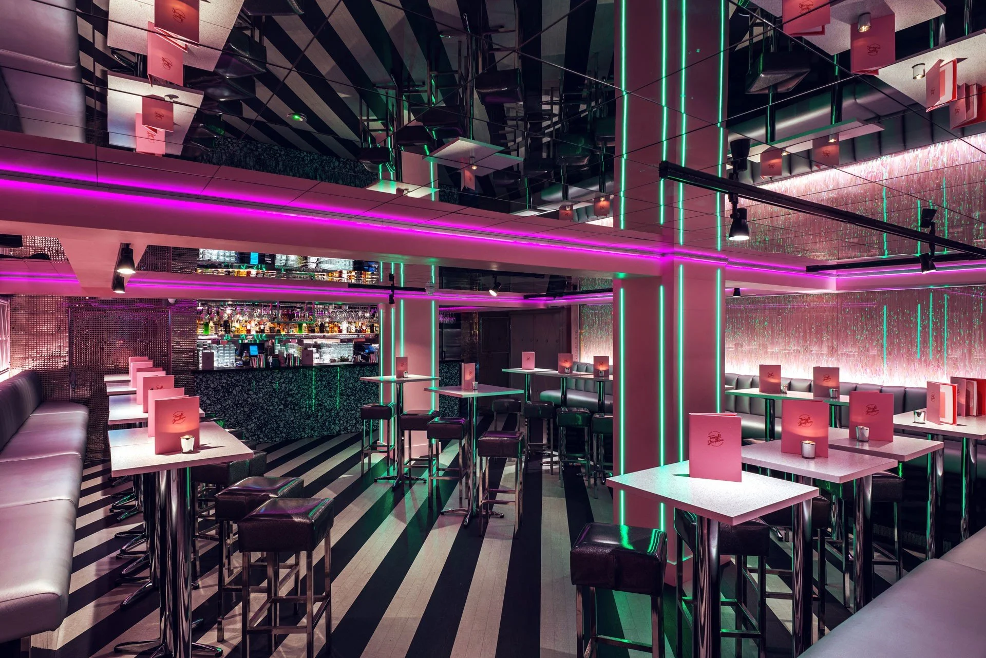 Interior of a modern cocktail bar with neon pink and green lighting, high tables with stools, and a terazzo bar counter in the background with bottles and glasses.