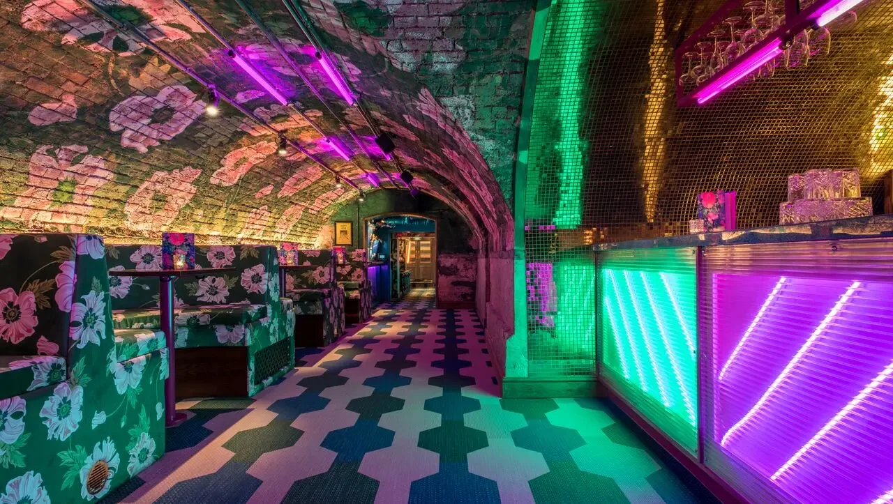 A vibrant nightclub with brick vaulted ceilings illuminated by purple and green neon lights, floral patterned booths, and a checkered floor.