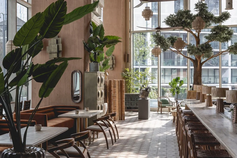 Modern interior of a cafe or restaurant with large windows, green plants, a large artificial tree, and natural sunlight.