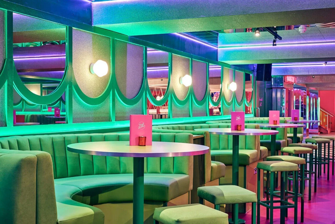 Colorful lounge with green booths, high tables, and pink menus, illuminated by green and purple lighting.