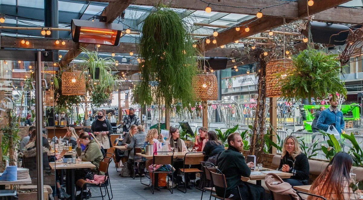 Indoor restaurant with wooden beams, hanging plants, and string lights, filled with patrons dining and talking.