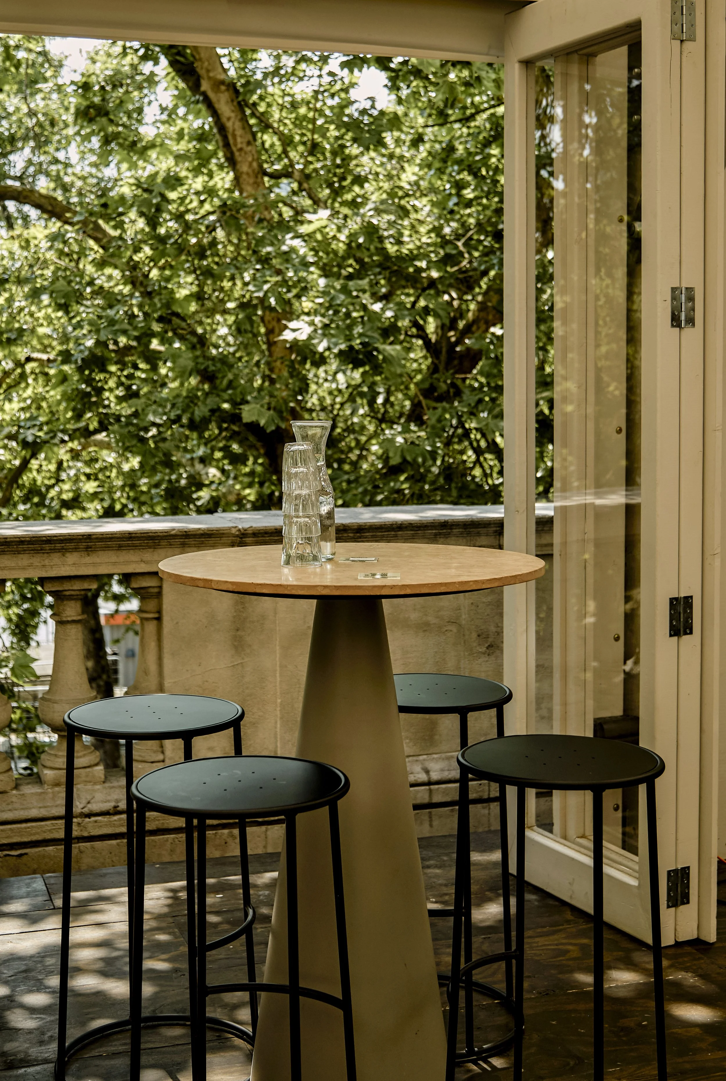 A small round table with glasses and a pitcher on top, surrounded by four barstools on a balcony with a green leafy view outside.