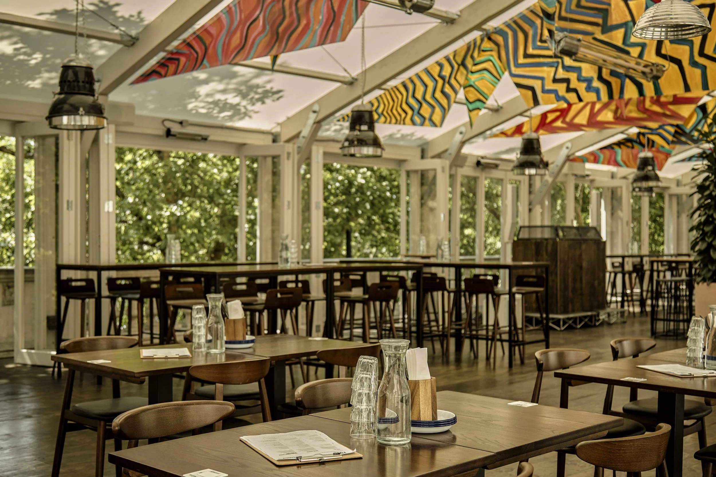 Empty restaurant interior with wooden tables, glasses, water bottles, colorful geometric-patterned fabric decorations hanging from ceiling, and large windows with greenery outside.