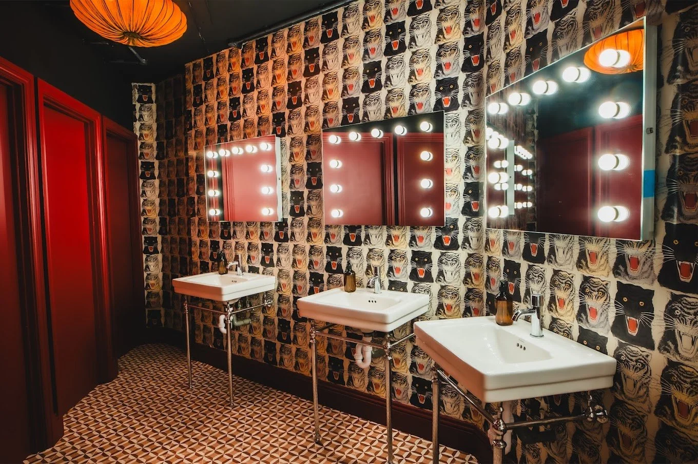 A bathroom with three sinks, large mirrors with lighting bulbs, tiger-themed wallpaper, red cabinets, and a geometric tile floor. There is a round orange paper lantern ceiling light.