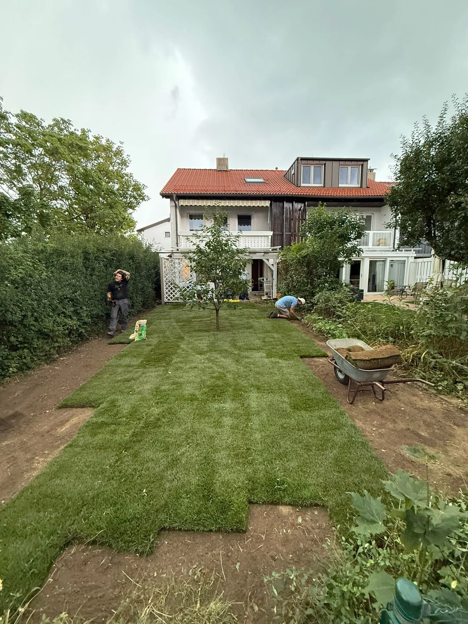 Zwei Personen pflanzen Rasen in einem Garten vor einem zweistöckigen Haus mit Balkon und Balkonüberdachung.