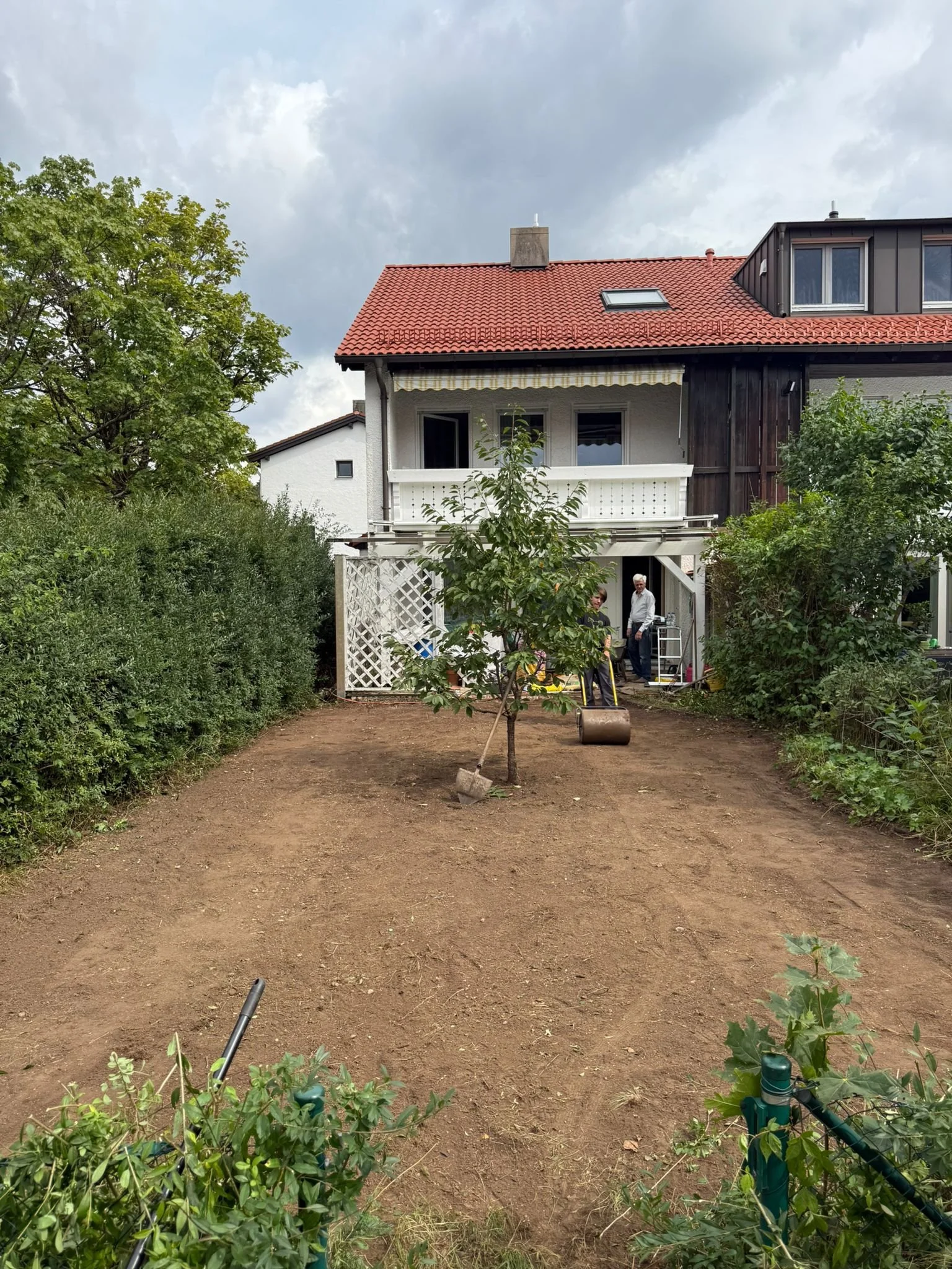 Ein Garten mit einem frisch umgegrabenen Erdteil, einem Baum in der Mitte, und zwei Personen im Hintergrund, die vor einem Haus stehen.