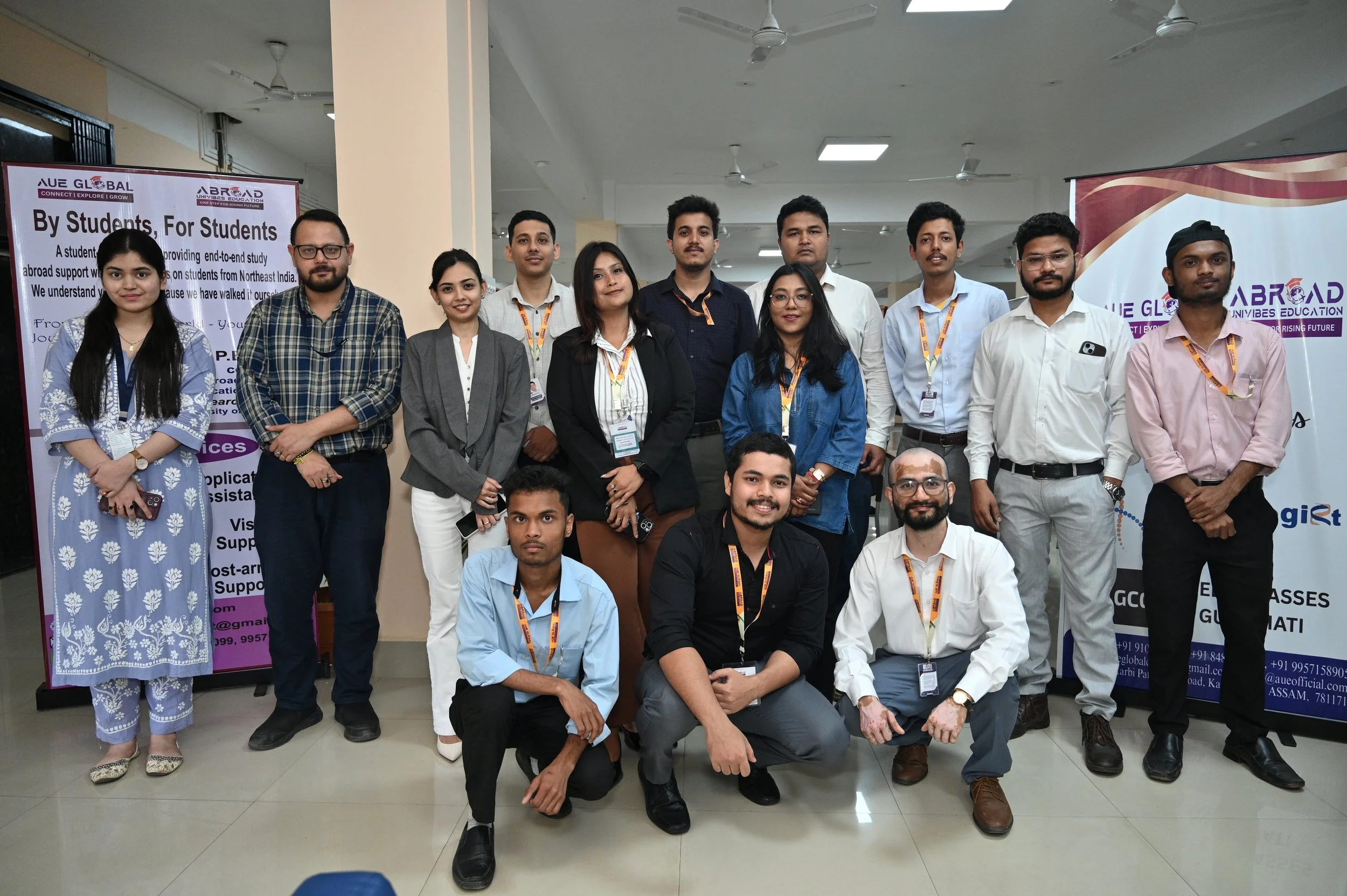 A group of young men and women posing together at an indoor event or conference. They are standing in front of banners with text about student support and education. The group is dressed in casual and formal attire, and some are wearing badges or lanyards.