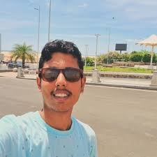 A young man with dark hair, wearing sunglasses and a light blue T-shirt, smiling in an outdoor setting with palm trees and a parking lot in the background.He is  the General Manager of Abroad Univibes Education( AUE Global ) Private Limited.