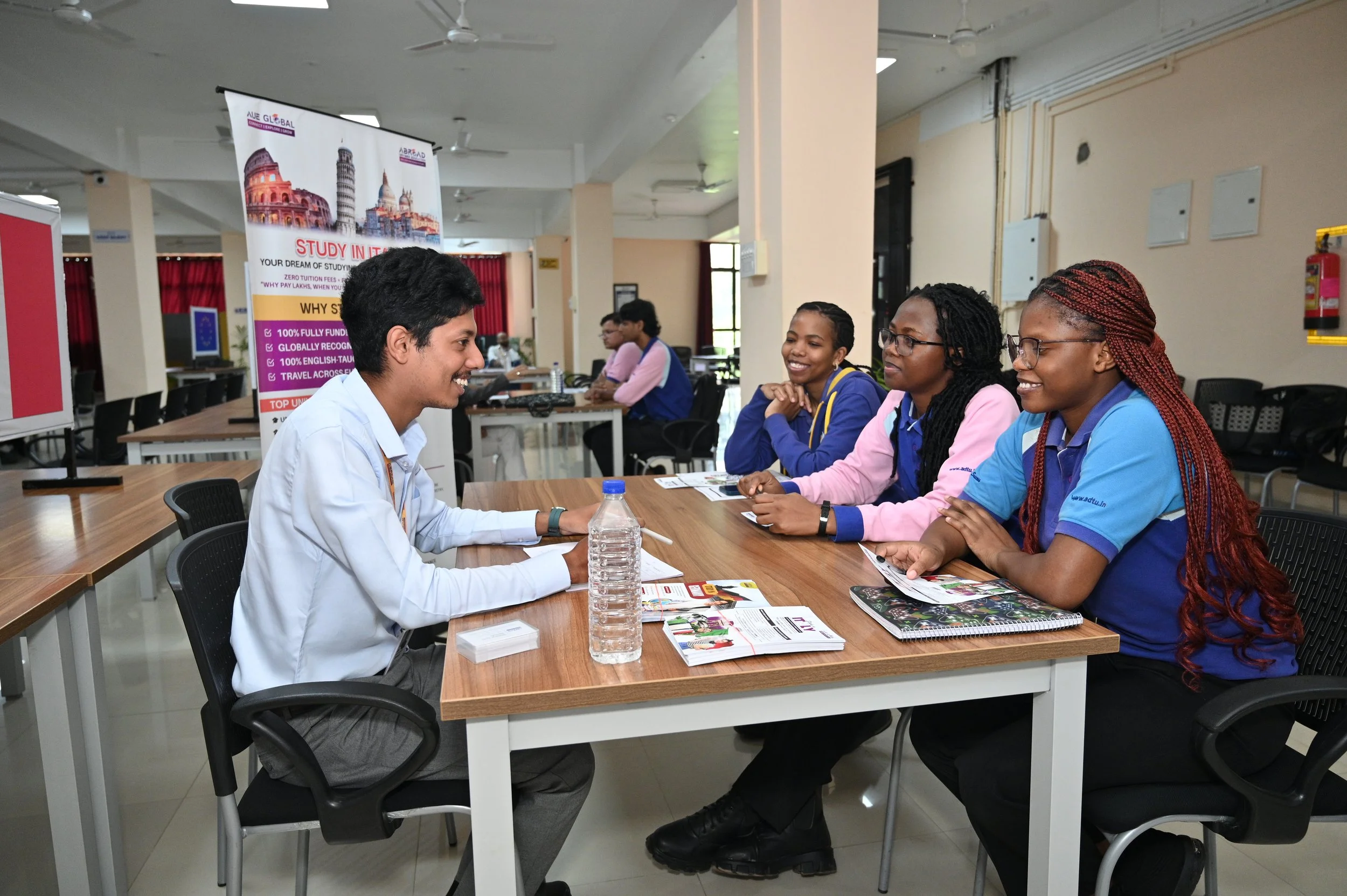 A group of students sitting at a table in a classroom or seminar room, engaged in a discussion, with a male student speaking and three female students listening attentively.