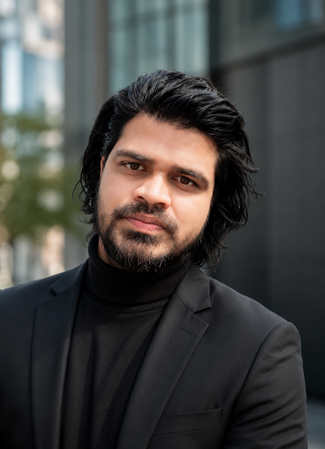 A man with black, wavy hair and a beard, wearing a black turtleneck and black blazer, standing outdoors in front of a modern glass building. He is a Founder of Abroad Univibes Education( AUE Global) Private Limited.