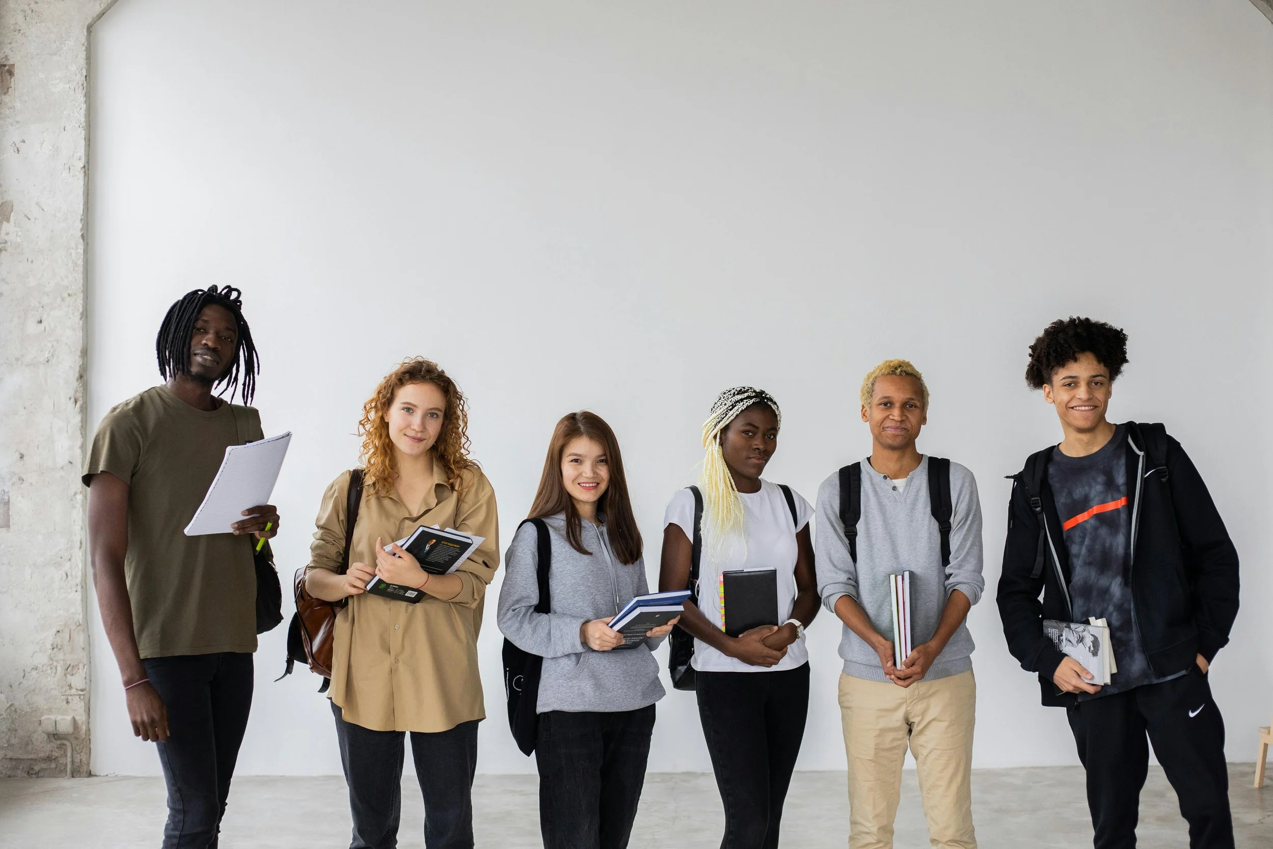 A diverse group of seven young students standing in a row against a plain white wall, holding books and notebooks, smiling and looking at the camera.