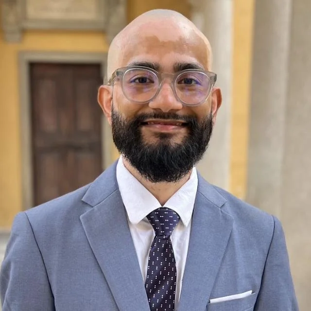 A bald man with a beard and glasses, dressed in a blue suit, white shirt, and a dark polka dot tie, smiling at the camera indoors with a blurred background. He is the Co-Founder of Abroad Univibes Education( AUE Global ) Private Limited.