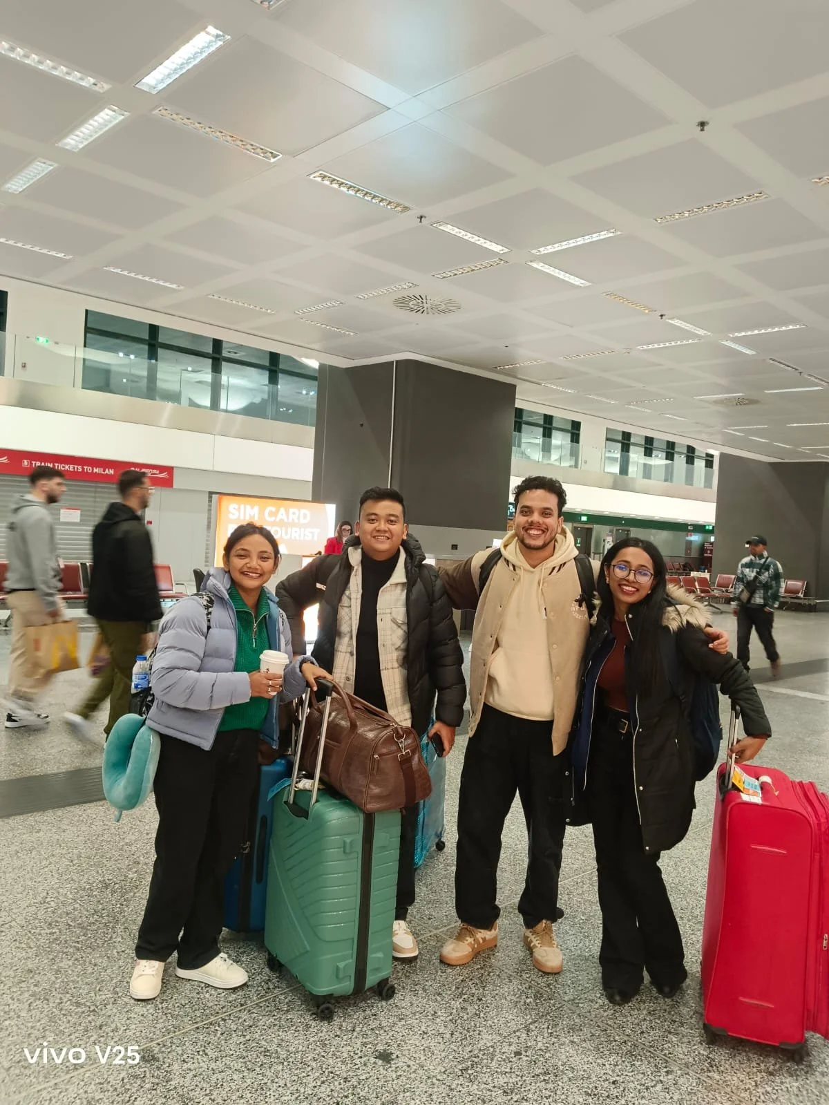A group of four young adults with suitcases and backpacks in an airport terminal, smiling at the camera.
