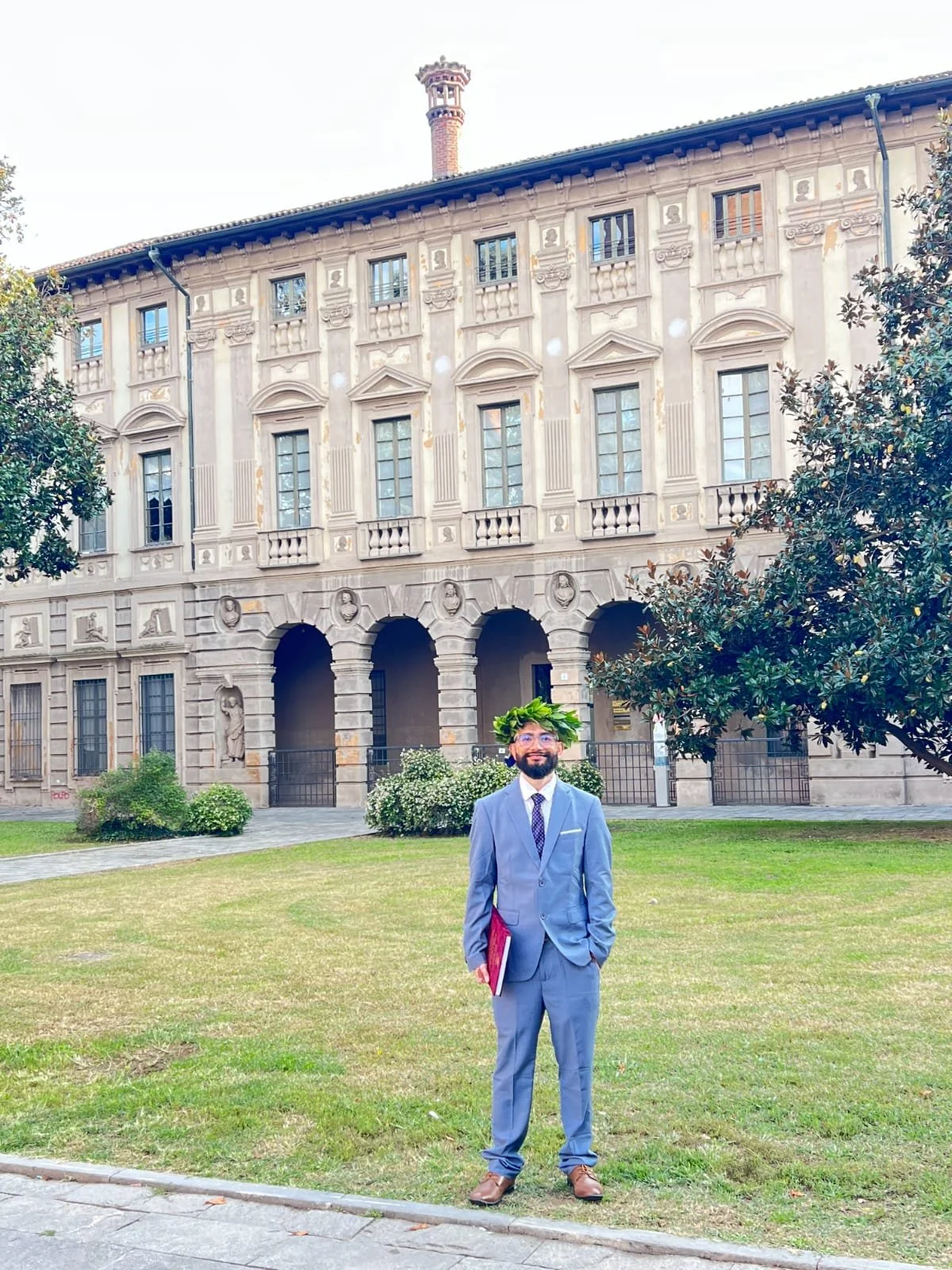 A man dressed in a gray suit with a plant crown on his head is standing on a lawn in front of a historic building with arched windows and decorative stonework, holding a red folder.