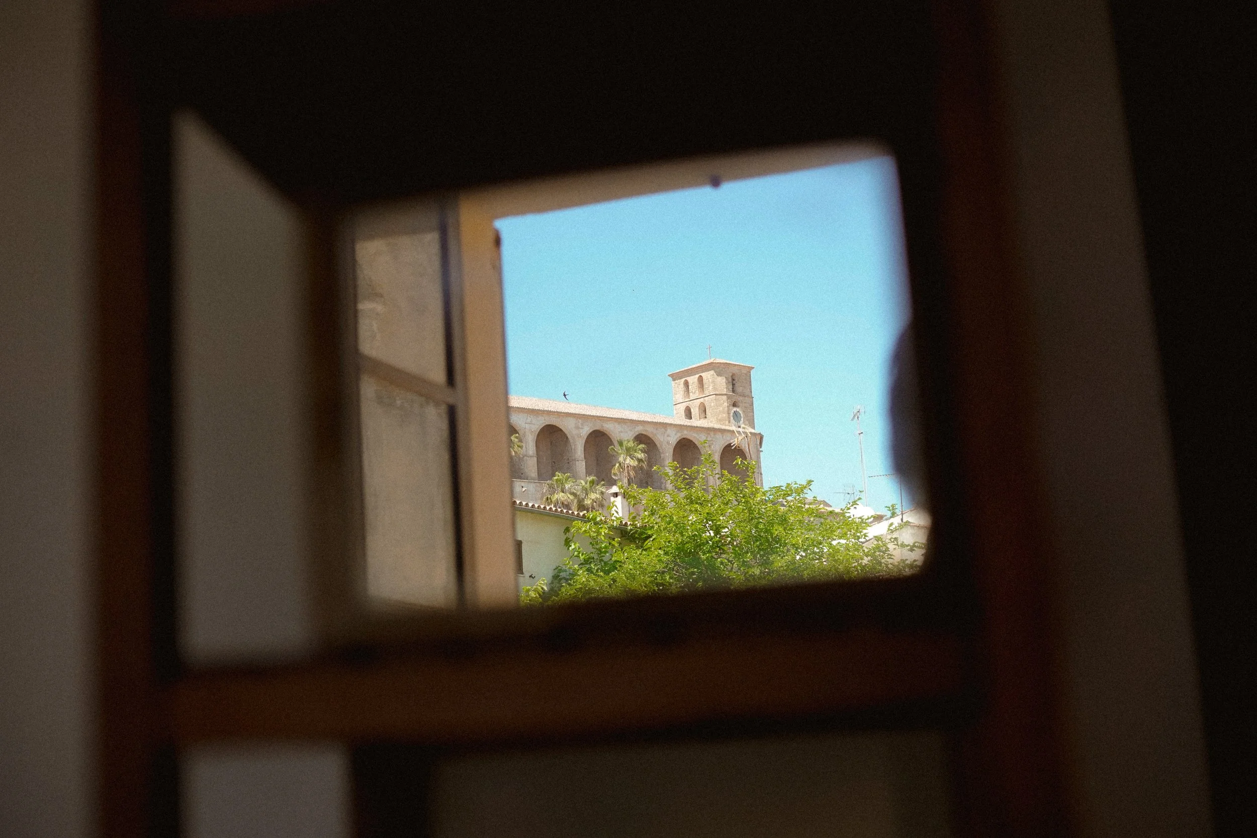 Blick durch ein kleines Fenster auf ein Kirchengebäude mit Kirchturm, auf blauem Himmel, umgeben von Bäumen.