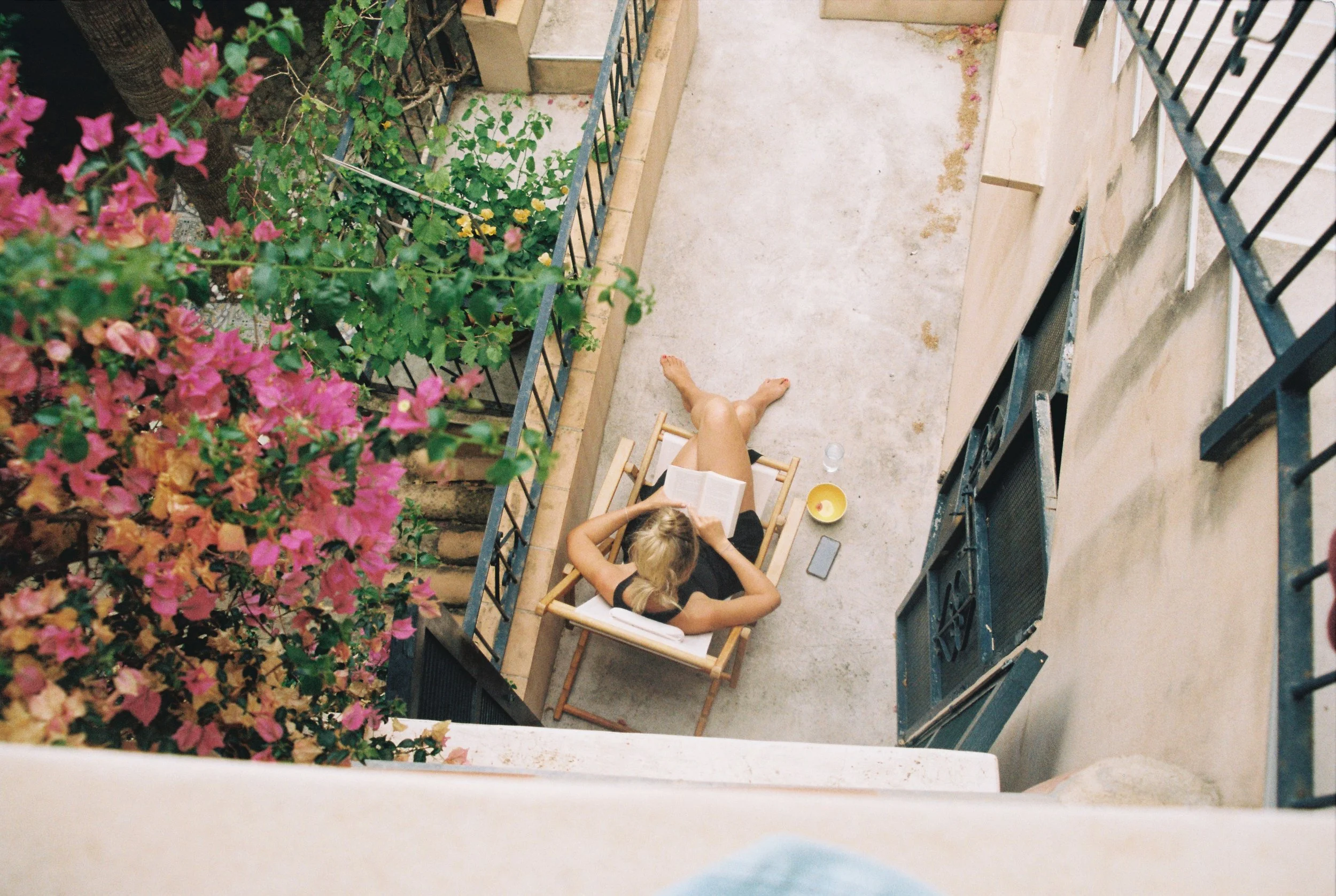 Zwei Frauen sitzen auf einem Balkon, lesen und trinken. Es gibt Pflanzen mit pinken Blumen auf der linken Seite und eine kleine Tasse auf dem Tisch.