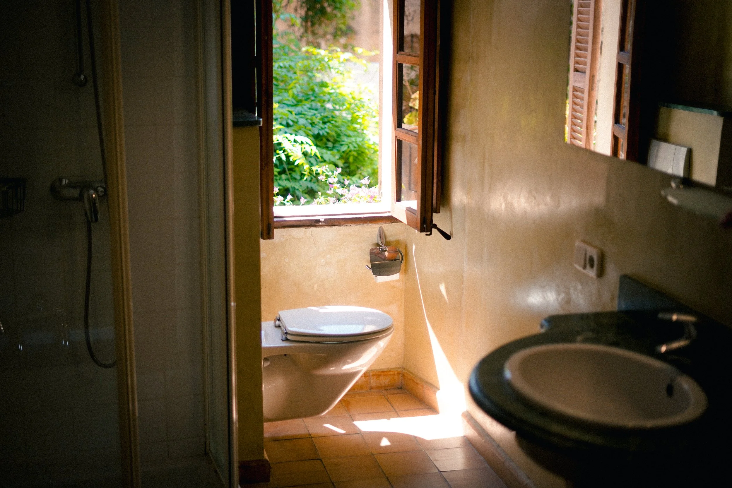 Ein Badezimmer mit geöffnetem Fenster, das nach draußen auf eine grüne Pflanzenlandschaft blickt, mit einer Toilette im Vordergrund.