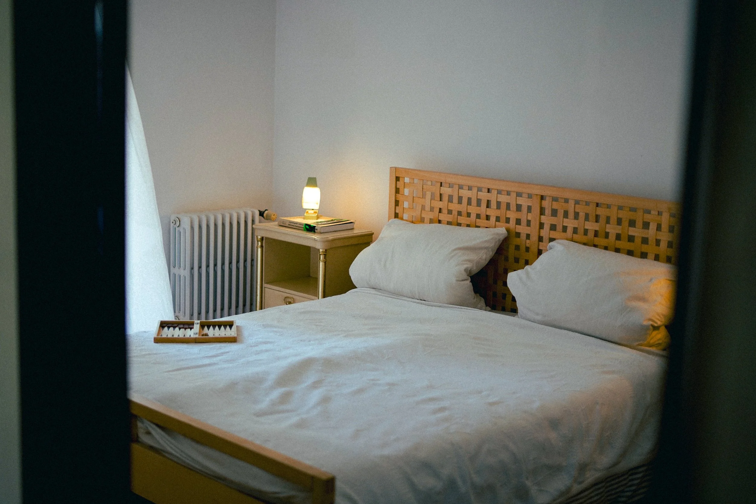 Bett mit weißen Bettwäsche und Kissen, Nachttisch mit Lampe, Büchern und einer Schachtel, im Hintergrund Heizkörper und eine weiße Wand.