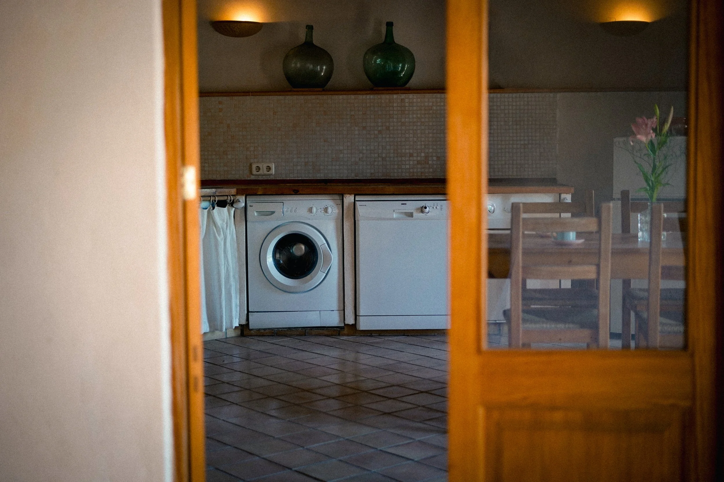 Blick durch eine gläserne Tür in eine Küche mit Waschmaschine, Spülmaschine, Esstisch und Stühlen, Dekorationen, wie Vasen und Blumen