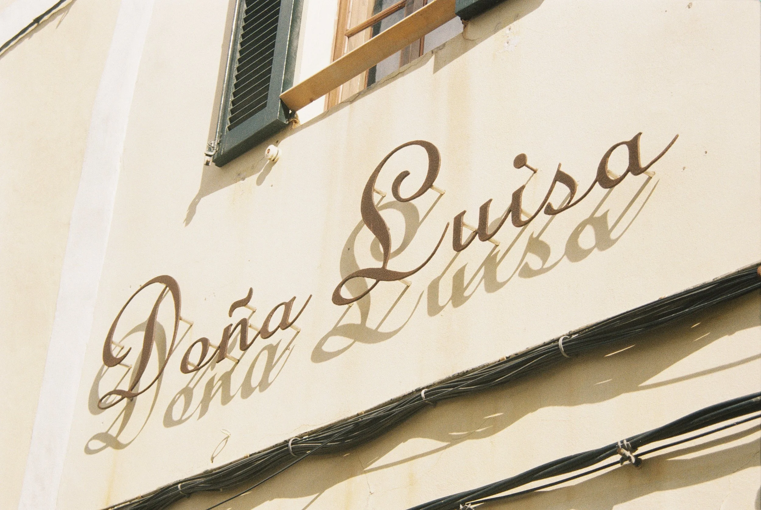 Die Aufnahme zeigt eine vertikale Außenfassade eines Gebäudes mit einer schwarzen Fensterläden und einem dekorativen Schriftzug in Schnörkelschrift, der wahrscheinlich den Namen eines Geschäfts oder Restaurants anzeigt.
