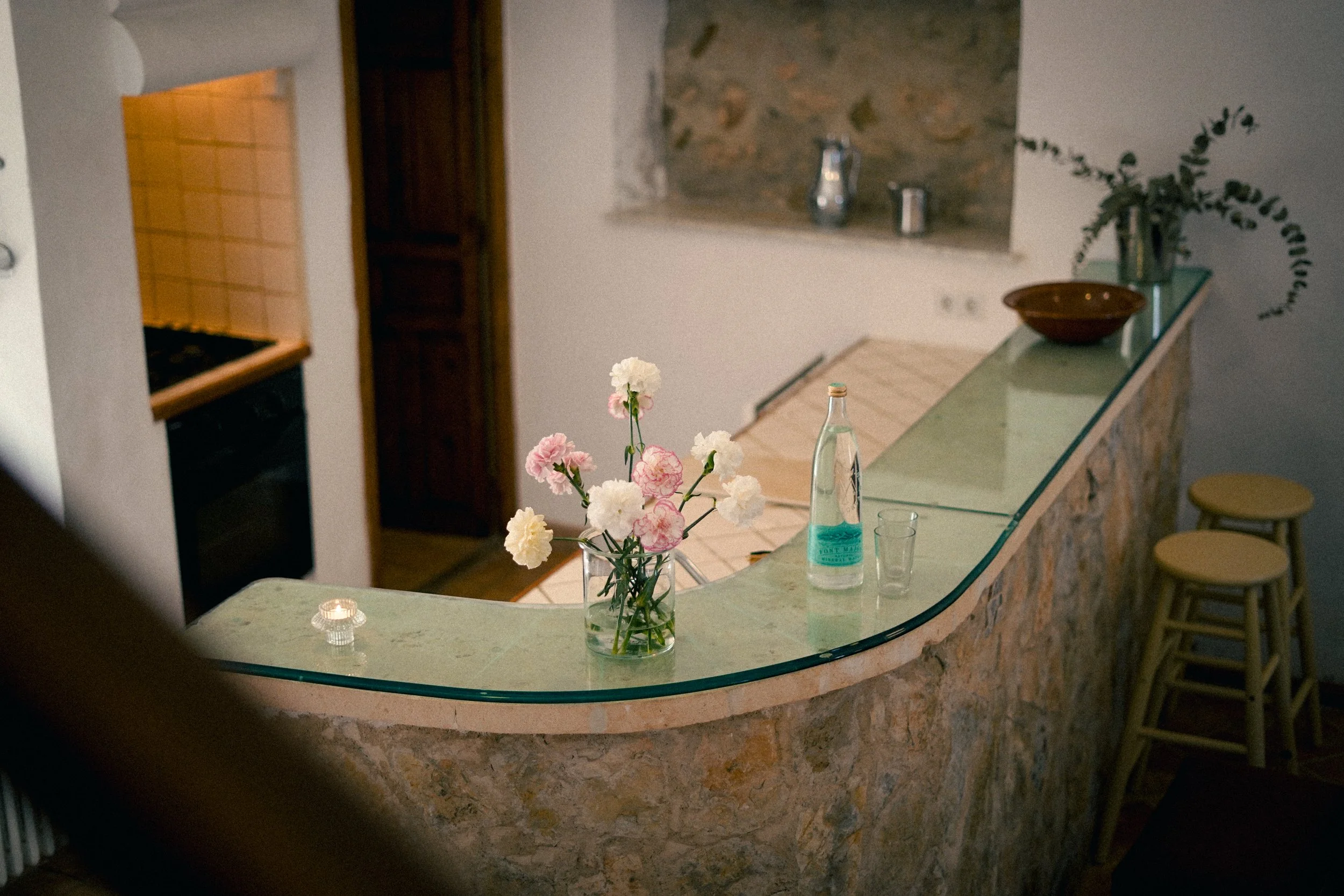 Ein Küchenbereich mit einem halbkreisförmigen Bartresen aus Stein, auf dem eine Vase mit weißen und rosafarbenen Blumen steht, sowie eine Wasserflasche und gläser. Im Hintergrund befinden sich eine Steinwand, eine Glasplatte mit Dekoration, und Barho