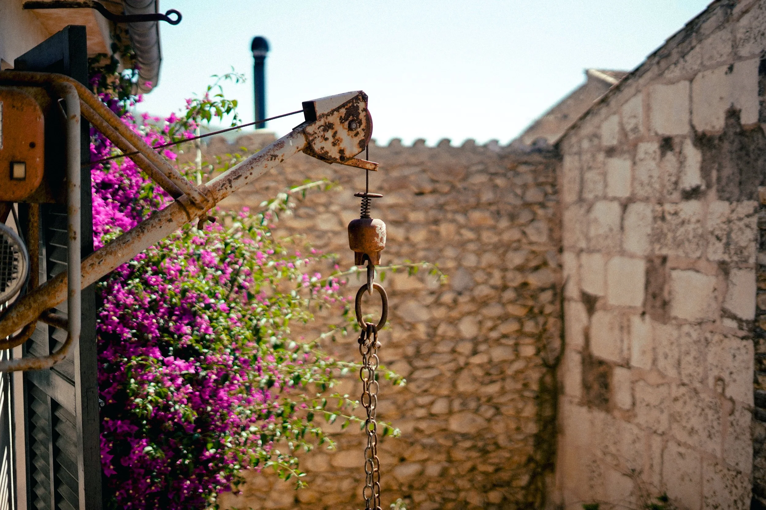 Ein verrostetes, altes Wäschewaschenetz an einer Kette hängt vor einer alten Steingmauer. Neben der Wand befindet sich eine rosa-blühende Pflanze.