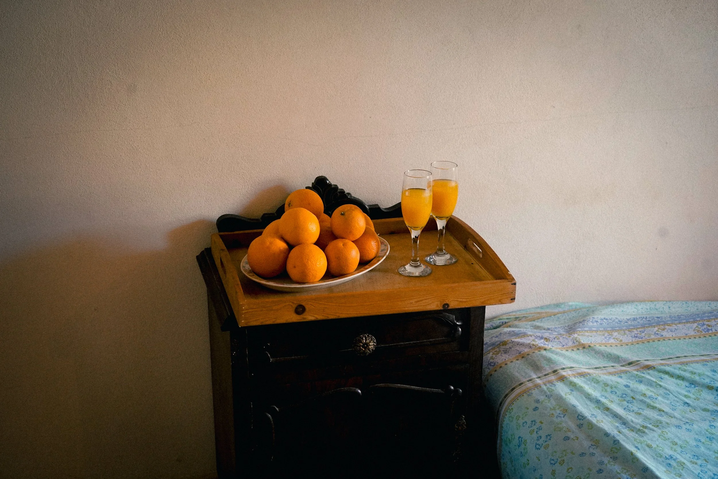 Ein Holztisch mit Orangen, Grapefruits und zwei Gläser Orangensaft vor einer schlichten weißen Wand, neben einem Bett mit gestreiftem Bettzeug.