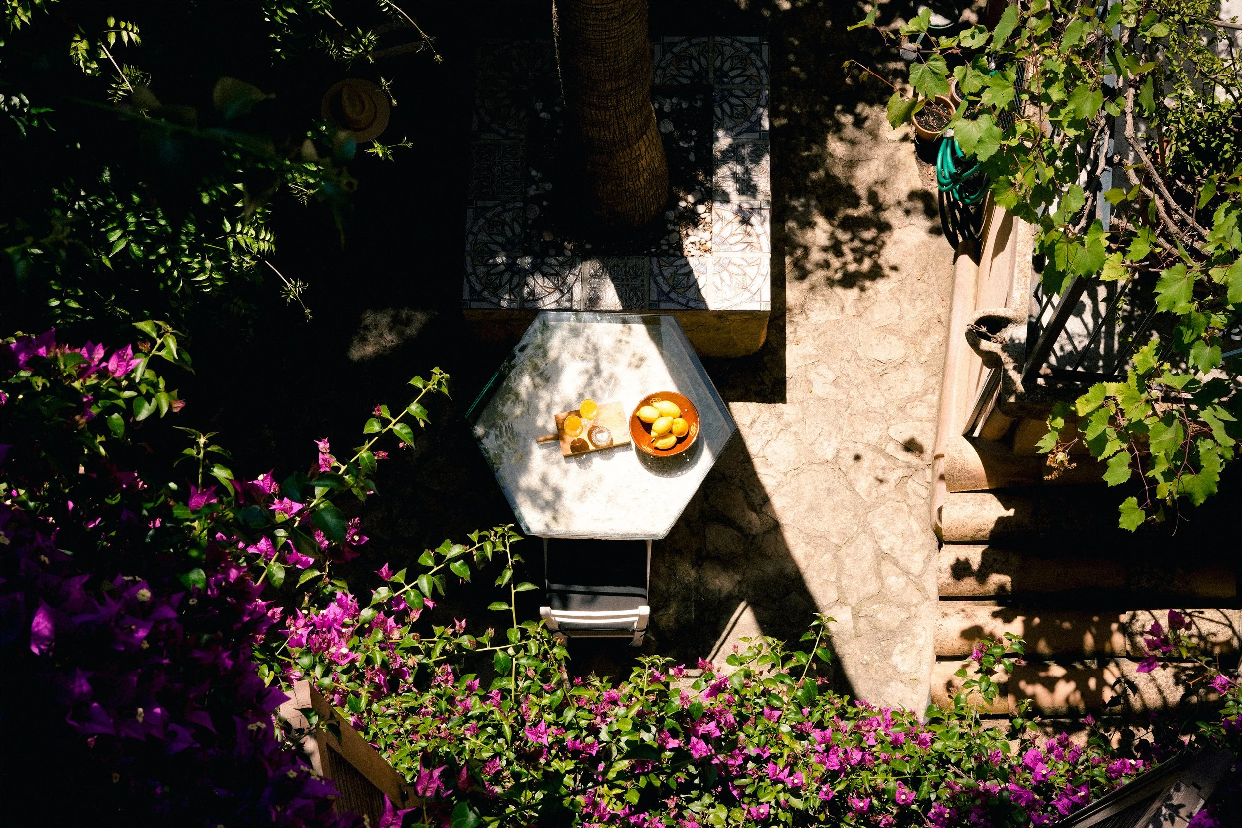 Blick auf einen kleinen Gartenbereich mit Pflanzen, Schatten und einem Steintisch mit Obst und Kerzen, umgeben von Blumentöpfen und einem Zaun.