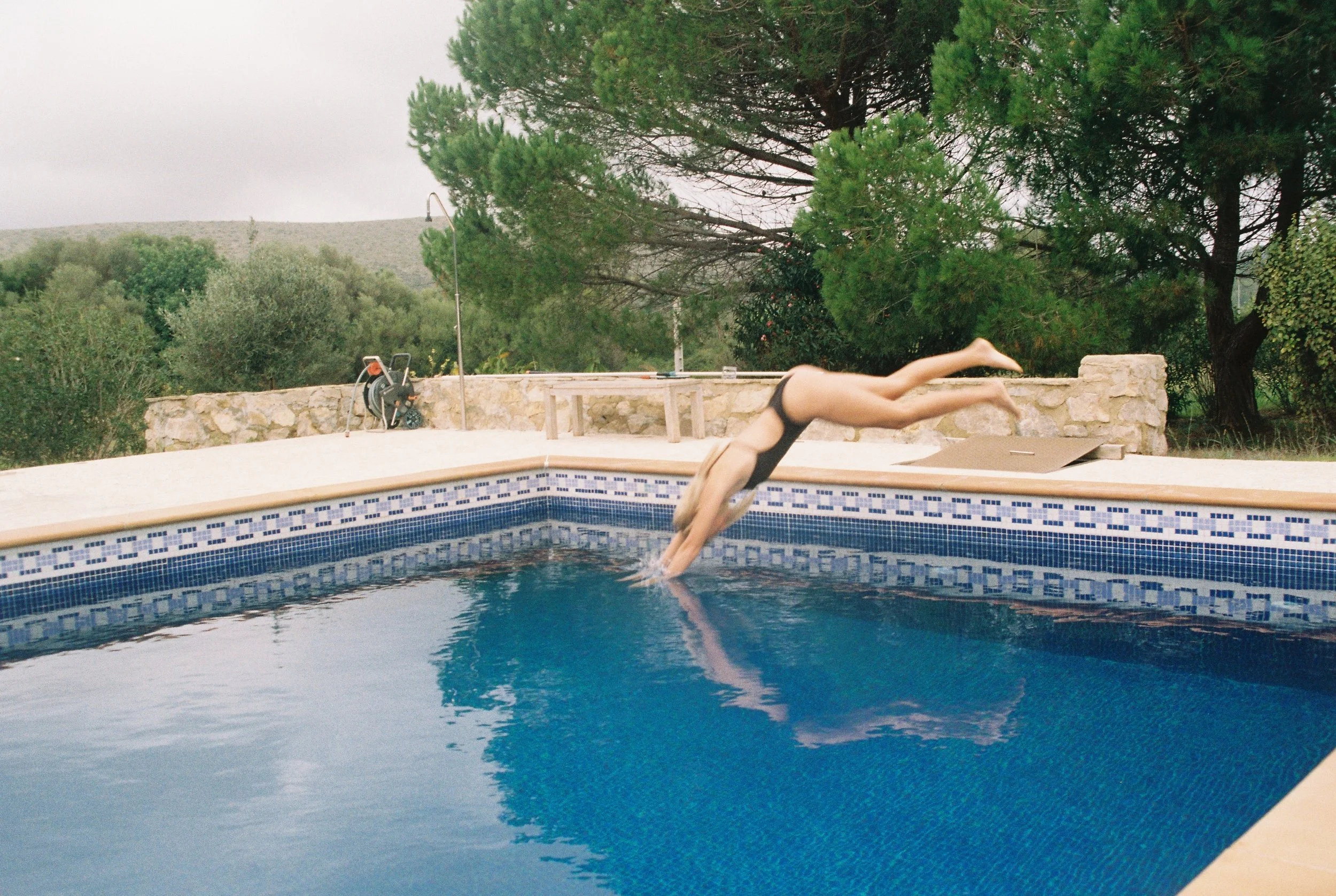 Eine Person springt kopfüber in einen Swimmingpool, umgeben von einer Steinmauer und grünen Bäumen, bei bewölktem Himmel.